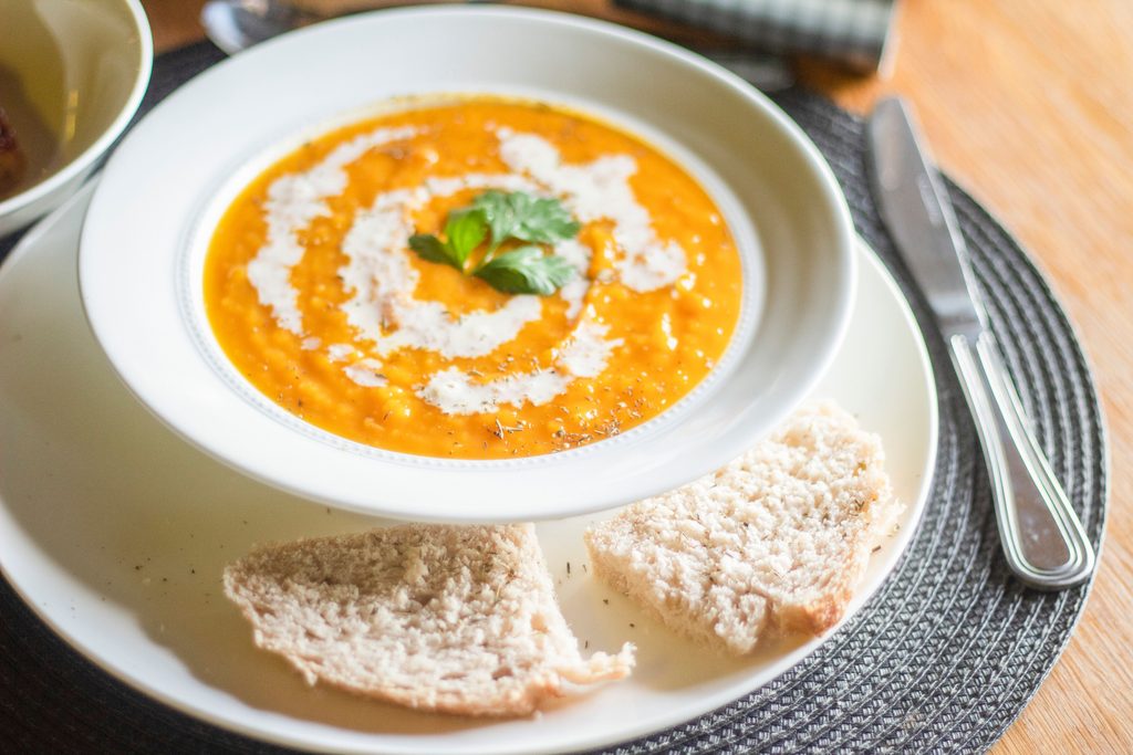 Bowl of roasted red pepper soup topped with cream and parsley next to bread. 
