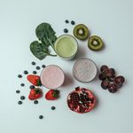 Smoothie ingredients including strawberries, kiwi, grapes, pomegranate, kale, and blueberries. Three smoothies in the middle of the smoothie ingredients.