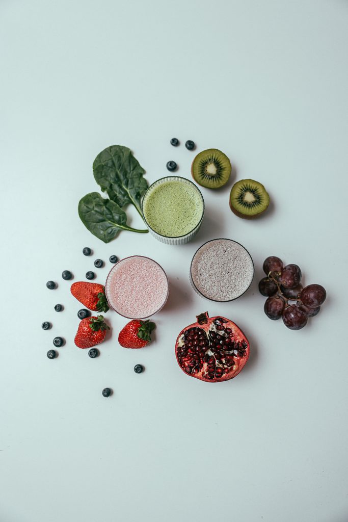 Smoothie ingredients arranged around three smoothies. Strawberry, kale, grape, kiwi, pomegranate. 